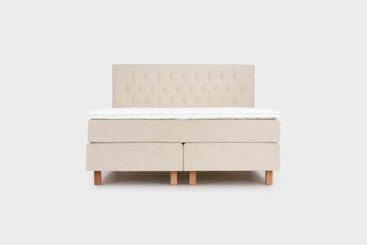 Vaalean beige jenkkisänky 180x210 cm tammen värisillä jaloilla ja vaalean beigellä sängynpäädyllä. Kuvattuna suoraan edestä päin vaaleanharmaata taustaa vasten.
