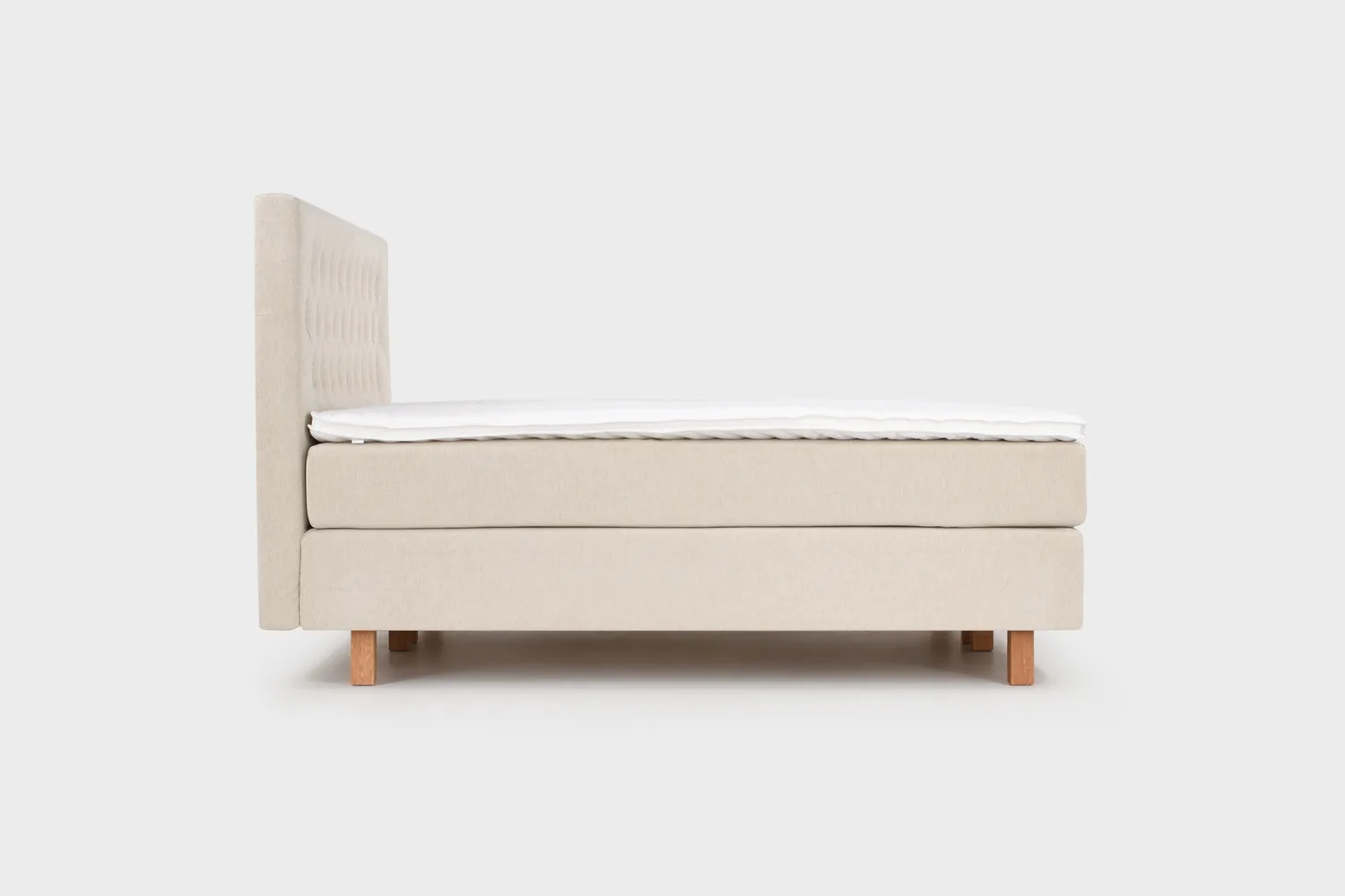 Vaalean beige jenkkisänky 180x210 cm tammen värisillä jaloilla ja vaalean beigellä sängynpäädyllä. Kuvattuna sivusta päin vaaleanharmaata taustaa vasten.