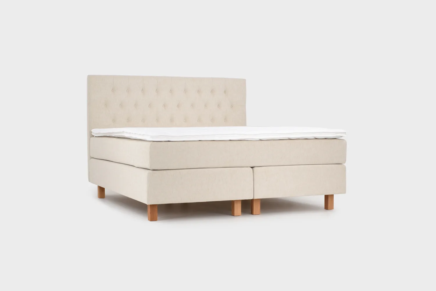 Vaalean beige jenkkisänky 180x210 cm tammen värisillä jaloilla ja vaalean beigellä sängynpäädyllä. Kuvattuna viistosti edestä päin vaaleanharmaata taustaa vasten.