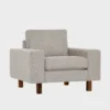 studio-xl-lepotuoli-100x90-das05sand-pahkina-finsoffat Beige kangasverhoiltu nojatuoli pähkinän värisillä jaloilla. Tuolissa on leveät käsinojat ja se on asetettu viistosti vaalean harmaata taustaa vasten.