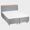 savoy-comfort-runkopatjasanky-180x200cm-das51-tammi-finsoffat Harmaa runkosänky 180x200 cm tammen värisillä jaloilla ja harmaalla sängynpäädyllä, jossa tammen värinen puukoristelista yläosassa. Kuvattuna viistosti vaaleanharmaata taustaa vasten.