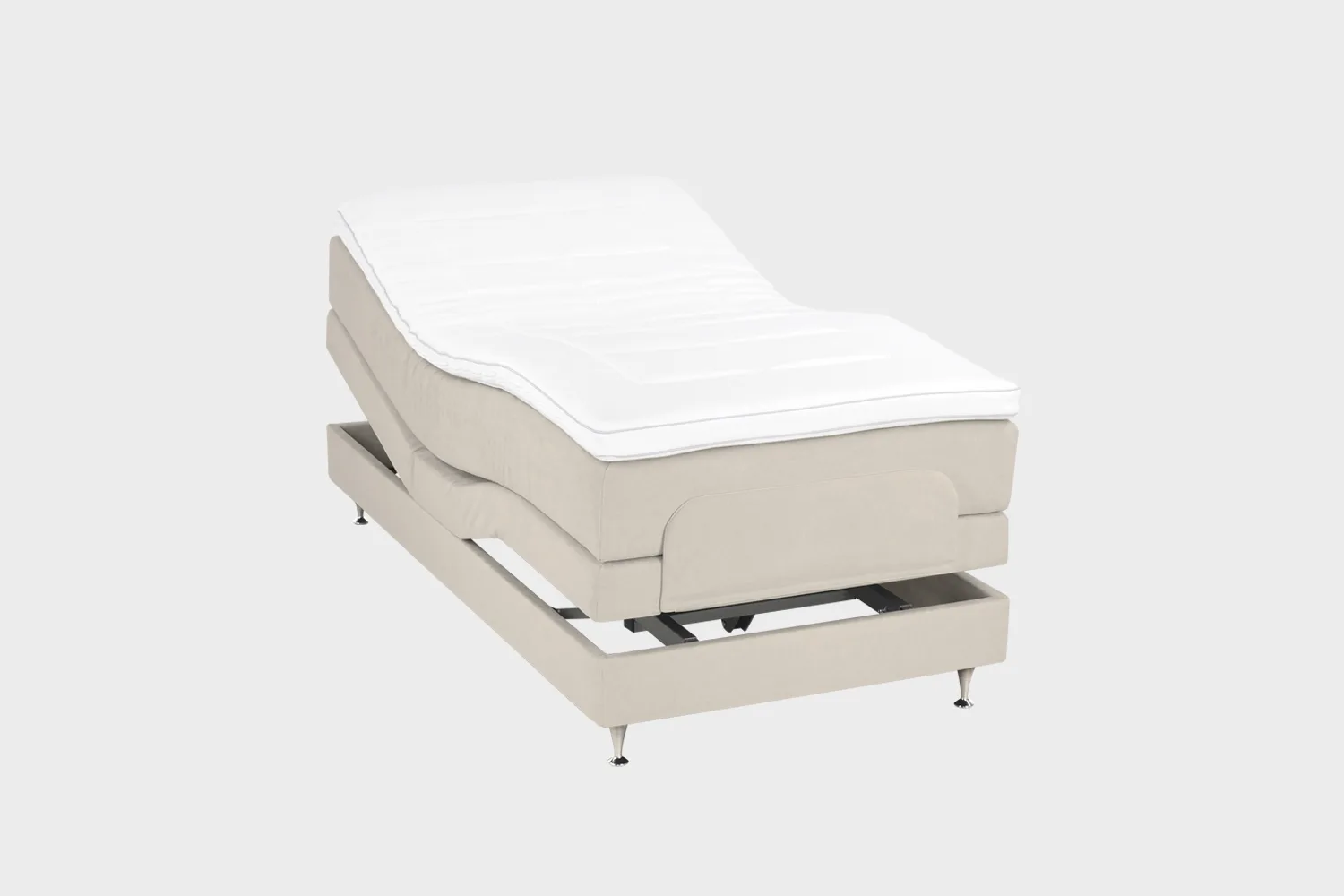 Vaalean beige moottorisänky 100x200 cm alumiinin värisillä jaloilla. Yhden hengen säätösänky on kuvattuna viistosti päädyt aavistuksen koholla vaaleaa taustaa vasten.